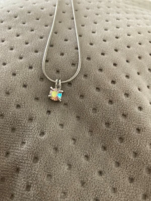 Silverfärgat halsband med regnbågssten - Ett stilrent halsband i silverfärg med en glittrande regnbågsskimrande ädelsten som hänge. Kedjan är tunn och smidig med justerbar längd. Perfekt för dig som gillar enkla men iögonfallande smycken.