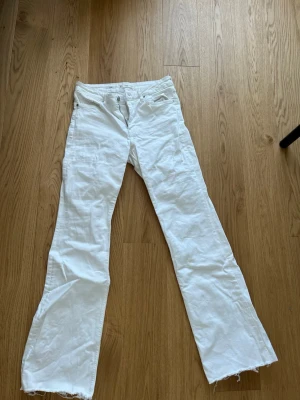 Högmidjade Vita bootcut jeans med rå kant från mango - Säljer ett par vita bootcut jeans med fransig, rå kant nedtill. Klassisk femficksmodell med normal midja och dragkedja. Jeansen är i ett mjukt bomullsmaterial och har en avslappnad passform. Perfekta för en trendig och avslappnad stil.