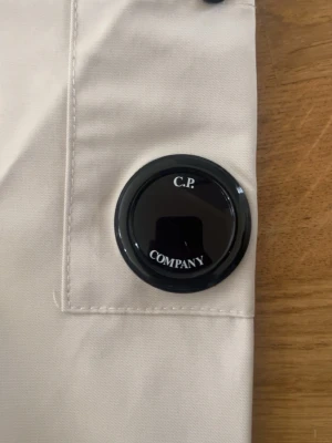 Beiga jeans från C.P. Company - Säljer ett par stilrena beiga jeans från C.P. Company med den ikoniska linsen på benfickan. Byxorna har en modern passform och är tillverkade i ett mjukt bomullsmaterial. Perfekta för dig som gillar streetwear och snygga detaljer.