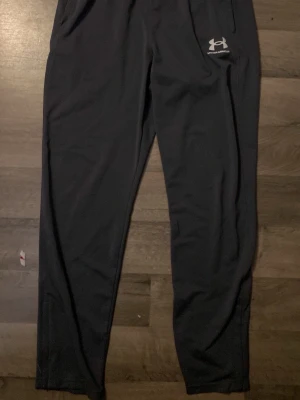 Mörk gråa träningsbyxor från Under Armour - Säljer ett par svarta träningsbyxor från Under Armour med logga på vänster sida. Byxorna har en normal passform och är tillverkade i ett mjukt syntetmaterial som passar perfekt för träning. Klassisk design utan mönster och med resår i midjan.