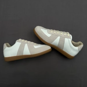 Vita och beige sneakers från Maison Margiella  - Stilrena sneakers från Maison Margiella i vitt och beige med ovandel i läder och mocka. Klassisk design med beige detaljer och snörning, samt en brun sula. Perfekta för en clean och avslappnad look.