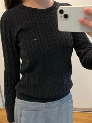 Svart kabelstickad tröja Tommy Hilfiger - Svart kabelstickad tröja från Tommy Hilfiger med rund halsringning och diskret logga på bröstet. Tröjan har långa ärmar och ribbade muddar vid ärmslut och nederkant. Perfekt för en stilren och klassisk look.