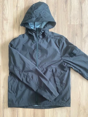 Svart vindjacka från Stone Island -  Äkta Svart vindjacka från Stone Island med huva och dragkedja framtill. Jackan har en ikonisk logotyp-patch på vänster ärm och är tillverkad i nylon och Tyvek-material. Perfekt för blåsiga dagar och har en modern, sportig look. Kan tänka mig att skicka båda badges vid rätt pris 