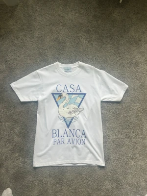 Vit Casa Blanca t-shirt med tryck - Vit t-shirt från Casablanca med stort grafiskt tryck framtill som visar en svan och texten 'Casa Blanca Par Avion' i blått. Klassisk passform och rund hals. Perfekt för dig som gillar stilrena men ändå unika plagg.