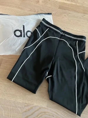 Svart träningsset från Alo - Snyggt svart träningsset från Alo med vita kontrastsömmar. Setet består av en sport-bh med breda axelband och ett par cykelshorts, båda i stretchigt funktionsmaterial. Perfekt för gymmet eller yoga.