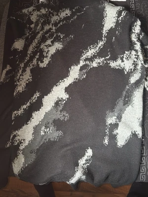 Svart stickad tröja med grått mönster - Cool svart stickad tröja med abstrakt grått mönster som ger en modern vibe. Tröjan har rund hals och är tillverkad i mjukt stickat material. Perfekt för dig som gillar unika plagg med streetkänsla.