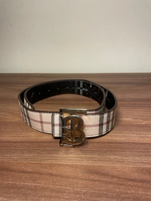 Randigt Burberry bälte med B-spänne - HELT NY!!! Säljer ett Burberry bälte med klassiskt randigt mönster och stort B-format metallspänne. Ett bälte som har två sidor i beige, brunt och rött och ett i ljusgrått och vitt. Perfekt accessoar för att lyfta din outfit med en ikonisk detalj. Lätt att byta sida
