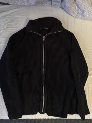 Hel Zip Tröja - Svart ribbad kofta från Lager 157 med hel dragkedja framtill och hög krage. Tröjan är långärmad och har en clean, minimalistisk look som passar perfekt till lager-på-lager. Materialet är stickat och känns mjukt mot huden. I princip oanvänd. Fraktas endast och köparen står för frakt.