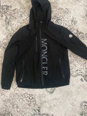  Moncler - Svart vindjacka från Moncler med huva, dragkedja framtill och två sidofickor med dragkedja. Stor Moncler-logga tryckt vertikalt på framsidan och klassisk Moncler-patch på ärmen. Jackan är tillverkad i ett lätt och smidigt material.