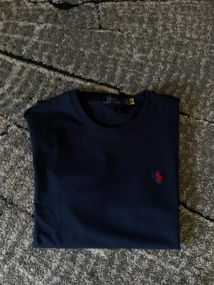 Mörkblå t-shirt från Polo Ralph Lauren - Klassisk mörkblå t-shirt från Polo Ralph Lauren med röd broderad logga på bröstet. T-shirten har rund halsringning och korta ärmar. Tillverkad i mjuk bomull för en skön känsla och enkel stil. Nypris runt 1000kr mitt pris 449kr aldrig använd!