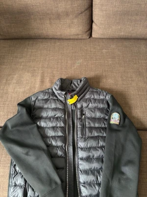 Svart dunjacka från Parajumpers - Säljer en svart dunjacka från Parajumpers med quiltad kropp och stickade ärmar. Jackan har en gul detalj vid kragen, bröstficka med dragkedja och Parajumpers-logga på ärmen. Perfekt för dig som vill ha både stil och funktion under vintern. QR koden funkar och leder till hemsidan 