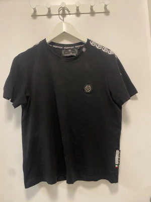 Svart t-shirt från Philipp Plein - Snygg svart t-shirt från Philipp Plein med logga på bröstet och vita logodetaljer längs axlarna. Rund halsringning och klassisk passform. Tillverkad i mjuk bomull för skön känsla. Perfekt för en trendig och avslappnad stil. . Passar perfekt till sommaren och till nästan alla outfits. Nästan helt ny, använt fåtal gånger. Kom med ett prisförslag om du är sugen, eller köp direkt såklart 😉 skickar samma dag! Står att de är M men passar mer S!