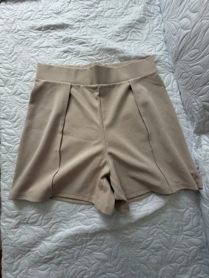 Beige högmidjade shorts från H&M - Stilrena beige shorts från H&M med hög midja och bred linning. Tillverkade i mjuk polyesterblandning med diskreta veck framtill för en snygg siluett. Perfekta för varma dagar och enkla att matcha med olika toppar.