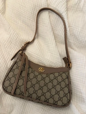 Gucci beige axelremsväska GG canvas - Säljer en beige axelremsväska från Gucci i klassisk GG canvas med bruna läderdetaljer och guldfärgad logga framtill. Väskan har justerbart axelband och dragkedja med lädertofs. Perfekt storlek för det viktigaste och riktigt trendig design. Tappat lite färg på metallen, se bilder.