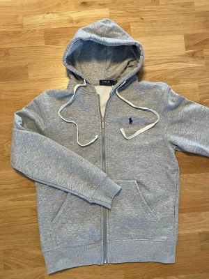 Grå zip hoodie från Polo Ralph Lauren - Snygg gråmelerad hoodie från Polo Ralph Lauren med dragkedja, vit dragsko i huvan och klassisk blå logga broderad på bröstet. Tillverkad i mjuk bomullsblandning och har två fickor framtill. Perfekt för en avslappnad och stilren look.