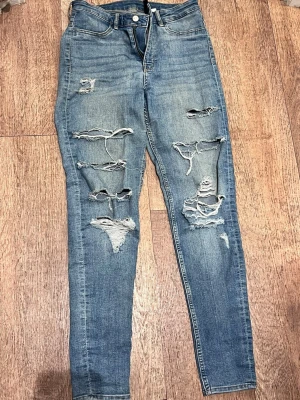 Blå slitna skinny jeans med hål - Säljer ett par blå skinny jeans med flera stora slitningar och hål framtill. Jeansen har klassisk femficksmodell, hög midja och dragkedja med knapp. Materialet är denim i bomull med lite stretch för skön passform.