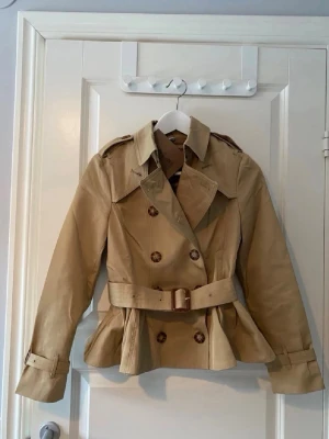 Kappa - Slutpris!!!Helt ny jacka med prislapp kvar. Klassisk trench-inspirerad modell i beige med skärp i midjan och dubbla knapprader. Bandet går att justera och ta av och på. Alla knappar går att knäppa av och på. Volang aktigt längst nere   Storlek står som 36, men jag skulle snarare säga att den passar 34 bäst. Högkvalitativ känsla och fint fall. Liten i storleken, den va för liten därav säljer jag den.
