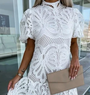 Vit spetsklänning med puffärm - Trendig vit klänning i spets med puffärmar och hög hals. Klänningen har ett detaljerat blommigt mönster och volang nedtill. Perfekt för sommarens alla tillfällen och ger en romantisk vibe.
