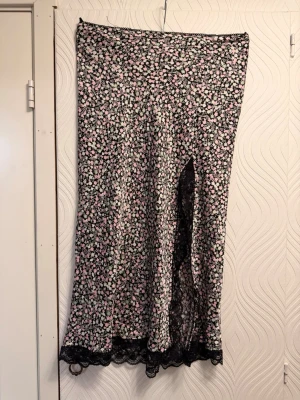Blommig långkjol med spets och slits - Säljer en svart långkjol med små rosa och vita blommor. Kjolen har en snygg slits framtill och spetsdetaljer längst ner. Perfekt för dig som gillar romantisk stil och vill ha något extra till garderoben. Dock en lös tråd från sömmen