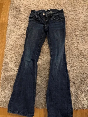Mörkblå bootcut jeans - Säljer ett par mörkblå bootcut jeans med klassisk femficksdesign och låg midja. Jeansen har en snygg tvättad look och vida ben som ger en retrovibe. Perfekta för dig som gillar en avslappnad men trendig stil.