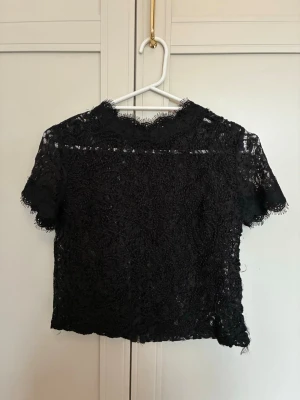 Svart spetsblus med dragkedja - Kort svart blus i spets med rund hals och korta ärmar. Baksidan har en synlig dragkedja och hela toppen är täckt av ett detaljerat spetsmönster. 