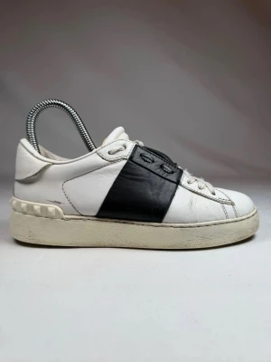 Valentino Garavani vita sneakers - Snygga vita sneakers från Valentino Garavani med svart läderdetalj över snörningen och ikoniska nitar på hälen. Skorna har en chunky sula och klassisk låg profil. Perfekta för dig som vill ha en stilren men ändå unik look.