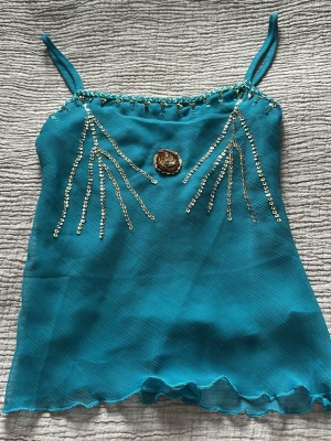 Turkos linnetopp med paljettdetaljer - Snygg turkos linnetopp med tunna axelband och dekorativa paljetter i guld framtill. Toppen har en vågig kant nertill och ett guldfärgat emblem i mitten. Perfekt för dig som vill sticka ut lite extra. Passar XS
