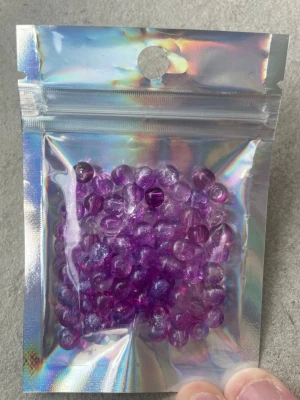Lila glaspärlor i holografisk påse - Ett paket med små, runda glaspärlor i klarlila nyans. Perfekta för att skapa egna smycken som armband, halsband eller örhängen. Pärlorna levereras i en holografisk zip-påse som ger extra wow-känsla.