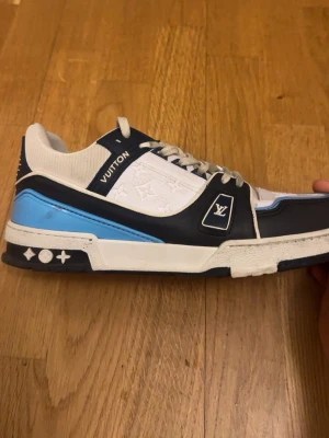 Louis Vuitton blå och vita sneakers - Snygga Louis Vuitton sneakers i vitt, blått och ljusblått med klassiska LV-loggor och monogramdetaljer på sidan. Skorna har lågt skaft, vita snören och en robust sula med ikoniska symboler. Perfekta för dig som vill sticka ut med exklusiv streetstyle.