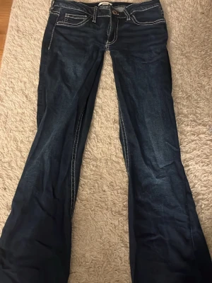 Mörkblå bootcut jeans från Gina tricot  - Säljer ett par mörkblå bootcut jeans från Gina tricot med kontrastsömmar och snygga detaljer på bakfickorna. Jeansen har låg midja, fem fickor och klassisk knappgylf. Perfekta för dig som gillar en avslappnad men trendig look.