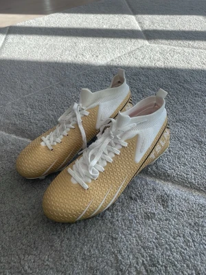 fotbollsskor  - Snygga fotbollsskor i guld och vitt från TSLN. Skorna har ett mönstrat syntetmaterial, vita snören och högt skaft för extra stöd. Sidorna pryds av vita detaljer och TSLN-logga. Perfekta för dig som vill sticka ut på planen. Storlek 38. Aldrig använda.