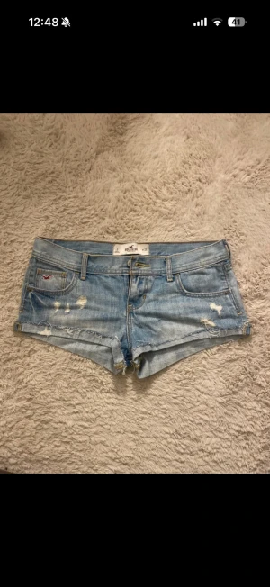 Ljusblå jeansshorts från Hollister - Säljer ett par ljusblå jeansshorts från Hollister med slitna detaljer och klassisk femficksdesign. Shortsen har låg midja och råa kanter för en avslappnad look. Perfekta till sommaren och lätta att matcha med olika toppar.
