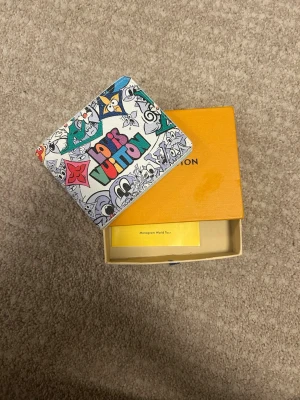 Louis Vuitton plånbok med graffiti - Färgglad plånbok från Louis Vuitton med graffitiinspirerat mönster och tecknade figurer. Plånboken har flera färger som blått, grönt, rött och rosa på vit bakgrund. Kommer i originalask. Perfekt för dig som gillar unika accessoarer.