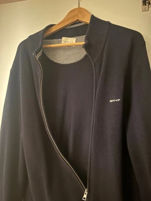 Mörkblå ziptröja från GANT - Snygg mörkblå ziptröja från GANT med diskret logga broderad på bröstet. Tröjan har hög krage, grovstickad struktur och dragkedja framtill. Perfekt för lager på lager och enkel att matcha med olika outfits.