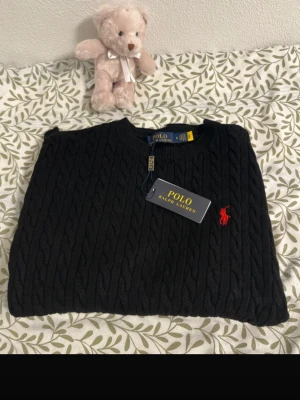 Svart kabelstickad tröja Polo Ralph Lauren - Kabelstickad svart tröja från Polo Ralph Lauren med rund halsringning och den klassiska röda logotypen broderad på bröstet. Tröjan har ribbade muddar vid hals, ärmslut och nederkant. Perfekt för dig som gillar stilrena och tidlösa plagg.