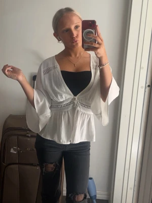 Vit omlott blus med vida ärmar - Säljer en luftig vit omlott blus med vida trekvartsärmar och dekorativa spetsdetaljer. Blusen har en v-ringad skärning och lätt rynkad midja som ger en avslappnad men chic look. Perfekt att styla över ett linne eller topp.