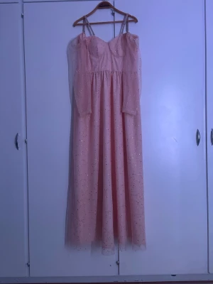 Ljusrosa glittrig långklänning - Säljer en romantisk ljusrosa långklänning med tunna axelband och glittriga detaljer över hela tyget. Klänningen har en lätt utsvängd kjol och en söt, feminin siluett. Perfekt för dig som vill sticka ut med lite extra glitter. Funkar till olika festsammanhang. Har bara provat och aldrig använt