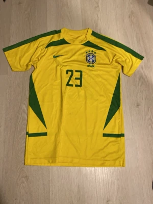 Brasilien fotbollströja Kaka #23 Nike - Gul Brasilien fotbollströja från Nike med gröna detaljer, nummer 23 och KAKA på ryggen. Tröjan har landslagets emblem på bröstet och är tillverkad i ett lätt, ventilerande material. Perfekt för dig som älskar fotboll och vill bära ditt favoritlag.