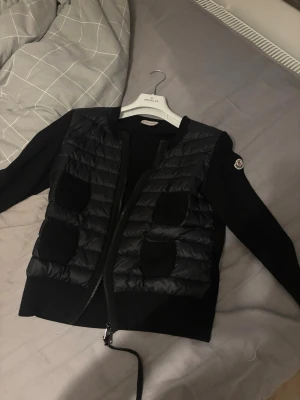 Svart cardiganjacka från Moncler - Snygg svart cardiganjacka från Moncler med quiltad front och fyra fickor. Jackan har Moncler-logga på ärmen och dragkedja framtill. Ärmar och rygg är i mjukt stickat material medan framsidan är vadderad för extra stil.