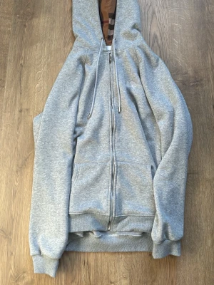 Grå zip hoodie från Burberry - Snygg grå zip hoodie från Burberry med klassiskt broderat riddarlogo på bröstet och huva med det ikoniska rutmönstret på luvan. Finns några små hål som är inuti hoodie men som man ej märker av 