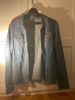 Blå jeansjacka från KappAhl - Klassisk blå jeansjacka från KappAhl i storlek 38. Jackan har två bröstfickor med knapp, knappar framtill och en rak passform. Perfekt att slänga över en hoodie eller t-shirt för en avslappnad look.