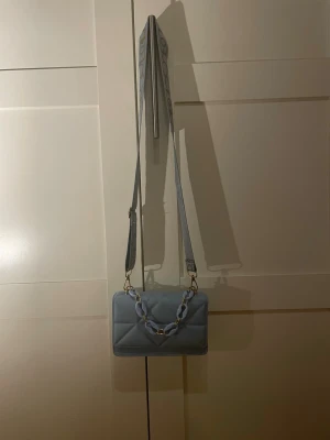 Ljusblå quiltad axelväska med kedja - Trendig ljusblå axelväska med quiltat mönster och bred justerbar axelrem med beige textdetaljer. Väskan har en chunky kedja framtill som handtag och är tillverkad i ett slätt, konstläderliknande material. Perfekt för dig som vill ha en cool och modern look.