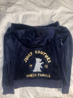 Mörkblå velour hoodie Juicy Couture - Mjuk och lyxig mörkblå hoodie i velour från Juicy Couture. Framsidan har dragkedja och fickor, baksidan har glittrigt tryck med hund, texten 'Juicy Couture', 'World Famous' och årtalet 1978 i guld. Perfekt för en chill och trendig look.