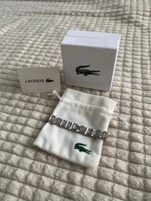 Silverfärgat armband från Lacoste - Snyggt armband i silverfärgat rostfritt stål från Lacoste med klassisk länkdesign och ingraverad krokodil på spännet. Kommer med originalpåse och ask med Lacoste-logga. Perfekt accessoar för dig som gillar stilrena detaljer.