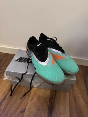 Nike Mercurial elite fotbollskor - Säljer ett par Nike Mercurial fotbollsskor i blåa med svarta och vita detaljer samt orange och gula accenter. Skorna har snörning, låg profil och ovandel i syntetmaterial för bra bollkontroll. Snygg och modern design som sticker ut på planen.
