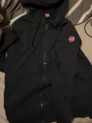 Mörkblå hoodie från Colmar med dragkedja - Säljer en mörkblå hoodie från Colmar med dragkedja och huva. Hoodien har Colmar-logga på bröstet och snörning i huvan. Perfekt för dig som gillar stilrena och klassiska plagg. Materialet känns mjukt och bekvämt.