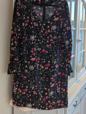 Blommig svart kortklänning med bälte - Svart kortklänning med färgglatt blommönster i rosa, gult, vitt och grönt. Klänningen har v-ringning, långa ärmar och ett matchande bälte i midjan. Perfekt för dig som gillar en feminin och lekfull stil.