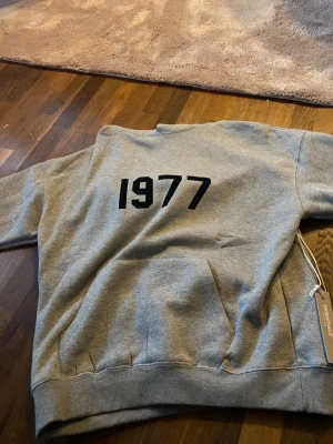 Grå Essentials hoodie med 1977-tryck - Säljer en grå hoodie från Essentials med svart '1977' tryckt på bröstet. Hoodien har en klassisk huva och magficka, samt ribbade muddar vid ärmar och nederkant. Perfekt för en chill och stilren look. Materialet känns mjukt och skönt mot huden.
