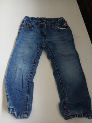 Blå jeans med broderi och resår - Säljer ett par blå jeans med resår i midjan och benslut. Jeansen har färgade sömmar och ett gulligt broderi med blommor och svamp på fickan. Klassisk femficksmodell i mjukt denimtyg, perfekta för en avslappnad look.