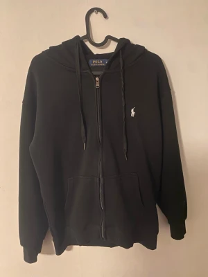 Svart zip hoodie från Polo Ralph Lauren - Säljer en svart hoodie med dragkedja från Polo Ralph Lauren. Klassisk design med huva och dragsnören, samt fickor framtill. Ikoniska vita Polo-loggan broderad på bröstet. Skön och mjuk bomullsblandning, perfekt för chill dagar. Storlek M men skulle mer säga att den är S